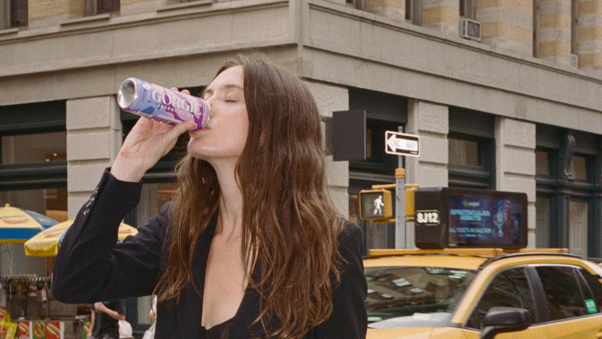 A woman in a black blazer and black shorts takes a drink from a purple can on a city street crosswalk, with a yellow taxi and a stone building in the background.